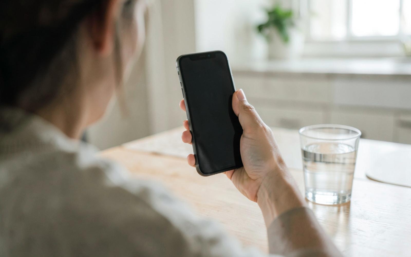 Person am hellen Küchentisch mit Telefon in der Hand und einem Glas Wasser