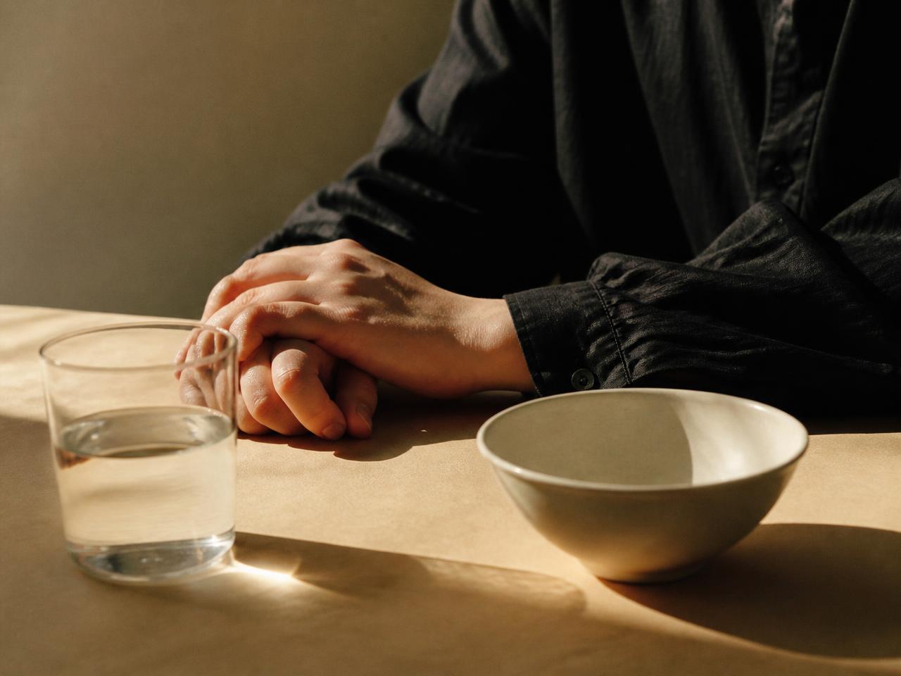 Hände ruhen neben einem Glas Wasser und einer schlichten Schale auf einem Tisch im Morgenlicht
